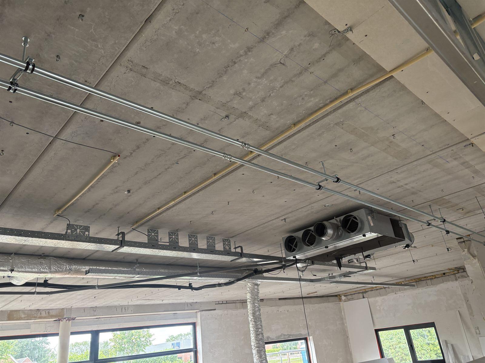 Unit boven het plafond gehangen in een bedrijfspand in aanbouw, leidingwerk op beugels langs het betonnen plafond